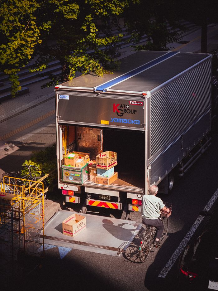 A delivery truck unloading boxes in a city street, with a bicyclist passing by on a sunny day.